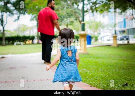 Asian 1-year-old toddler è occupato a piedi con suo padre in un parco tropicale di mattina. Esplorazione e la paternità del concetto. Foto Stock