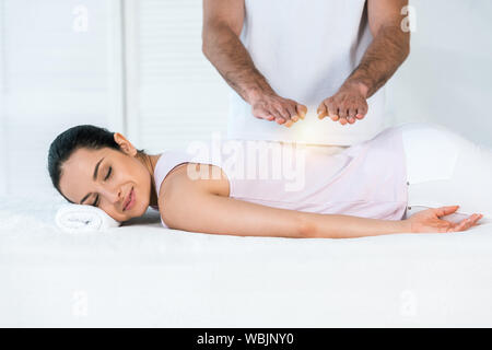 Vista ritagliata di guaritore di mettere sopra le mani donna attraente con gli occhi chiusi sdraiato sul lettino da massaggio Foto Stock