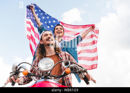 Basso angolo vista della bellissima ragazza a cavallo con il giovane uomo su scooter rosso e tenendo bandiera americana Foto Stock