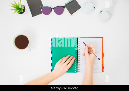 Flatlay con mani femminili la scrittura in un blocco note. Area di lavoro di Office. Sfondo bianco e la tazza di caffè accanto a. Foto Stock