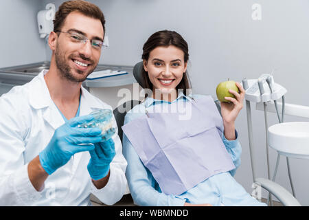 Messa a fuoco selettiva di allegro dentista in bicchieri che mantiene i denti modello vicino a donna felice con mela verde Foto Stock