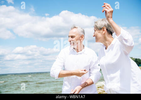Sorridenti coppia senior in camice bianco gesticolando in spiaggia sotto il cielo blu Foto Stock