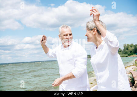 Sorridenti coppia senior in camice bianco guardando ogni altro e gesticolando in spiaggia sotto il cielo blu Foto Stock