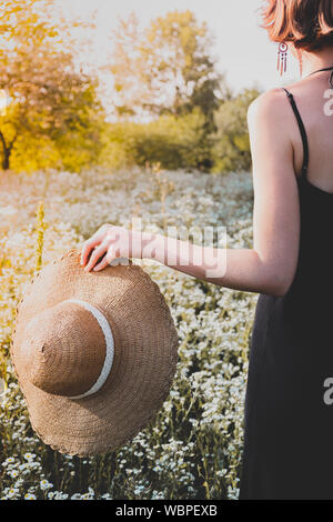 Persona di amare e di apprezzare la natura del concetto. Cappello di paglia in mani femminili su un grazioso prato rurale pieno di fiori selvatici Foto Stock