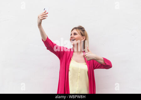 Come selfie tempo! Ritratto di felice attraente blogger donna in rosa camicetta in piedi, tenendo il telefono e rendendo selfie e mostra Thumbs up. indoor, s Foto Stock