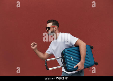 Giovani viaggiatori. Vista laterale del bello uomo barbuto in occhiali da sole tenendo il suo bagaglio mentre in piedi contro la parete rossa all'esterno. Concetto di viaggio Foto Stock