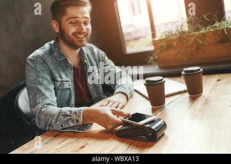 Ritratto di giovane uomo il pagamento fattura tramite lo smartphone utilizzando la tecnologia NFC in cafe. Takeaway due tazze di caffè sul tavolo. Inquadratura orizzontale Foto Stock