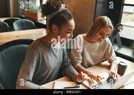 Ritratto di due multirazziale giovane femmina amici navigando in internet insieme su un laptop come si siedono in una caffetteria gustando una tazza di caffè. Horizo Foto Stock