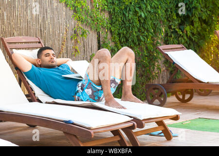 Ritratto di bello barbuto giovane adulto uomo freelancer in maglietta blu e pantaloncini giacente su cozysunbed con laptop e dormire dopo una giornata di duro lavoro. Lifestyl Foto Stock