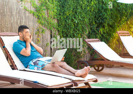 Ritratto di stanchi bello barbuto giovane studente adulto uomo in blu t-shirt e shorts sdraiato sul letto con il computer portatile sul bordo della piscina e istruzione online, co Foto Stock