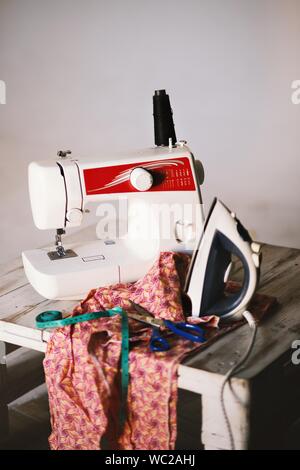 colpo di primo piano di una macchina da cucire bianca, tessile, forbici, metro a nastro e ferro da stiro su un tavolo Foto Stock