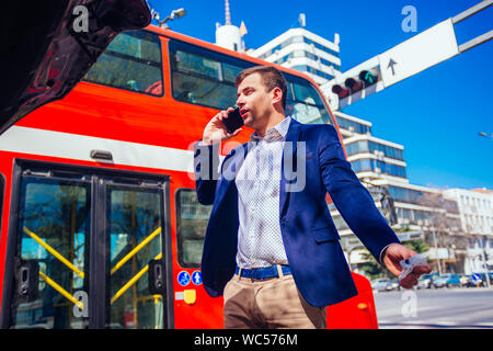 Giovane imprenditore sta parlando sul suo telefono cellulare mentre guardando sotto il cofano sollevato della sua vettura. Foto Stock