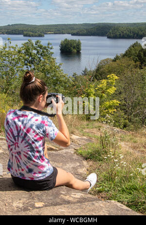 Una ragazza adolescente di scattare foto a un belvedere al di sopra di un lago e a una foresta in estate a Muskoka. Foto Stock