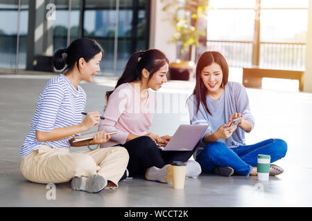 Gruppo di giovani smart studenti asiatici che ridere rendendo note guardando laptop e discutendo seduti sul pavimento esterno Foto Stock