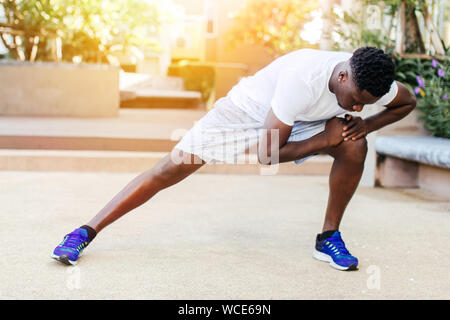 Athletic African American uomo in blu sneakers facendo esercizio di stretching mentre la formazione nel parco sulla giornata di sole Foto Stock