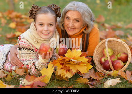 Graziosa bambina con la nonna giacente su erba, vicino a cesto in vimini pieno di mele nel parco autunnali Foto Stock