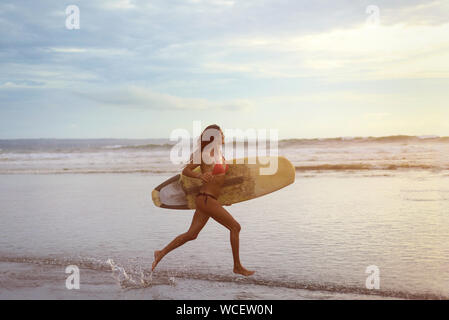 Una giovane donna con il bianco navigare nelle sue mani che corre lungo la sponda dell'oceano al tramonto. Gli spruzzi di acqua. A tutta lunghezza foto Foto Stock