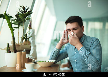 Giovane uomo bello soffia il naso mentre insalata di mangiare e di bere il caffè per il pranzo durante la pausa al cafe vicino ufficio sembra malato. Foto Stock