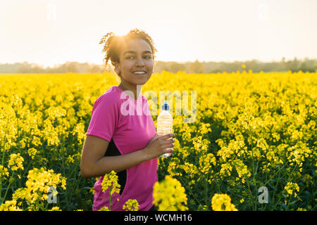 Outdoor ritratto della bella felice razza mista americano africano ragazza adolescente di sesso femminile donna giovane atleta runner acqua potabile da una bottiglia in un fiel Foto Stock