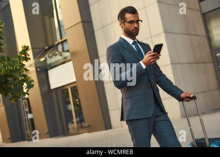 Pronto ad aiutare ovunque. Giovane e bello uomo barbuto in tuta valigetta e guardando il suo smartphone permanente, mentre all'esterno. In viaggio. Il concetto di business Foto Stock