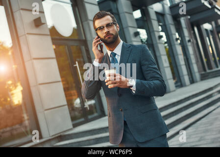Importante parlare. Giovane e bello barbuto imprenditore nel usura formale parla al telefono e bicchiere di contenimento di caffè permanente, mentre all'esterno. Il concetto di business Foto Stock