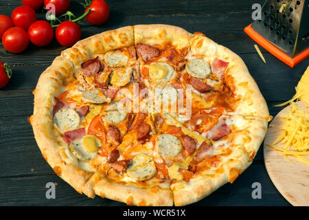 Tagliare la pizza, su fette, con salsiccia, pomodoro e formaggio. Sul tavolo di legno vi sono anche i pomodori e il formaggio grattugiato. Close-up. Foto Stock