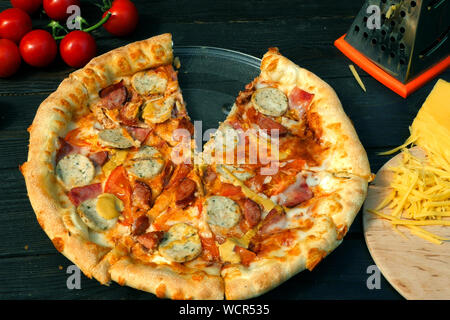 Tagliare la pizza, su fette, con salsiccia, pomodoro e formaggio. Sul tavolo di legno vi sono anche i pomodori e il formaggio grattugiato. Close-up. Foto Stock