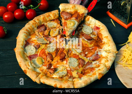 Tagliare la pizza, su fette, con salsiccia, pomodoro e formaggio. Sul tavolo di legno vi sono anche i pomodori e il formaggio grattugiato. Close-up. Foto Stock