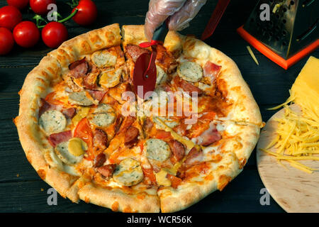 Tagliare la pizza, su fette, con salsiccia, pomodoro e formaggio, con uno speciale coltello - coltello ruota . Sul tavolo di legno vi sono anche i pomodori e grattugiato chee Foto Stock