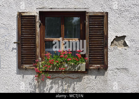 Vecchia finestra spiovente di una vecchia casa colonica nella campagna circondata da vecchia facciata con intonaco fatiscente e una scatola di gerani di fronte al w Foto Stock