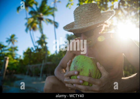 Uomo che indossa una paglia cappello per il sole seduto con una noce di cocco verde retroilluminati da sole tropicale in una remota isola beach Foto Stock
