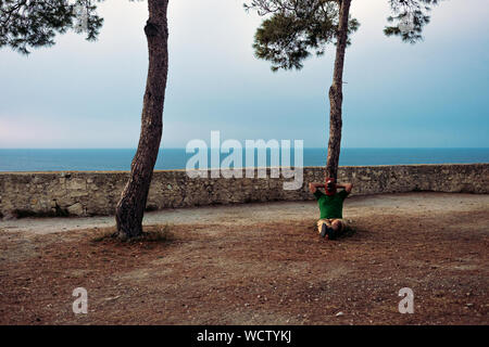Uomo con una testa di cavallo maschera rilassa seduto sotto un italiano in legno di cembro con il mare in background Foto Stock