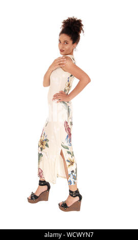 Una bellissima giovane multi-razziale donna in piedi in una lunga estate vestito e tacchi in profilo, con una mano sulla sua anca, con il suo nero capelli ricci in un panino, iso Foto Stock