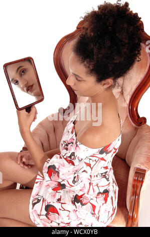 Bella multi-razziale donna che guarda a se stessa in uno specchio, seduta in abiti estivi e ricci capelli neri, isolato per sfondo bianco Foto Stock