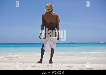 Giovane uomo che indossa un cappello di paglia mentre in piedi su una spiaggia che guarda al mare. Foto Stock