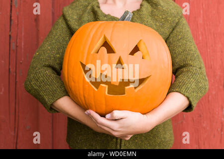 Close-up di giovane donna in possesso di una zucca jack-o-lantern all aperto in autunno con un granaio rosso in background Foto Stock