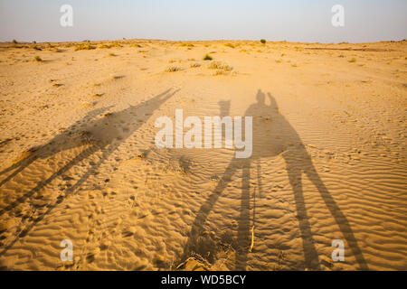 L ombra di un uomo a dorso di un cammello e scattare una foto della sua ombra sul deserto arido terreno. Deserto di Thar, Rajashan, India. Foto Stock