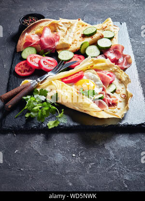 Deliziosa crepes con uovo fritto, prosciutto a fette, freschi Pomodori e cetrioli ripieni su una pietra nera vassoio con ingredienti, vista verticale dall'alto, Foto Stock
