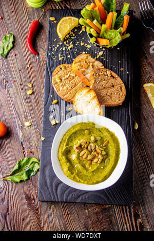 Hummus con spinaci, avocado e semi di zucca in una ciotola su una tavola di legno e bruschetta, orientale e cucina araba , Orientamento verticale Foto Stock