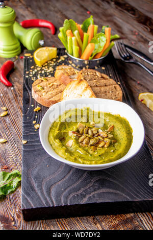 Hummus con spinaci, avocado e semi di zucca in una ciotola su una tavola di legno e bruschetta, orientale e cucina araba , Orientamento verticale Foto Stock