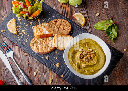 Hummus con spinaci, avocado e semi di zucca in una ciotola su una tavola di legno e bruschetta, cucina orientale, orientamento orizzontale, vista dall'alto Foto Stock