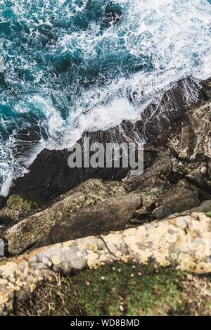 Angolo verticale alto girato dalla cima della scogliera vicino al mare con le onde che colpiscono la roccia Foto Stock