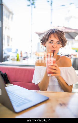 Lifestyle Moda Ritratto di giovane ragazza piuttosto bere il frullato al cafe' all'aperto. Sorridente e godendo di pasto. Indossando Elegante cappello di paglia, occhiali da sole Foto Stock