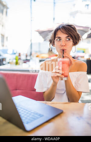 Lifestyle Moda Ritratto di giovane ragazza piuttosto bere il frullato al cafe' all'aperto. Sorridente e godendo di pasto. Indossando Elegante cappello di paglia, occhiali da sole Foto Stock