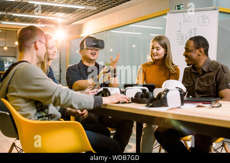 Il lavoro di squadra cercando occhiali per realtà virtuale per il lavoro in ufficio creativo. Giovane uomo asiatico utilizzando la nuova tecnologia occhiali vr mentre i suoi colleghi parlare sitti Foto Stock