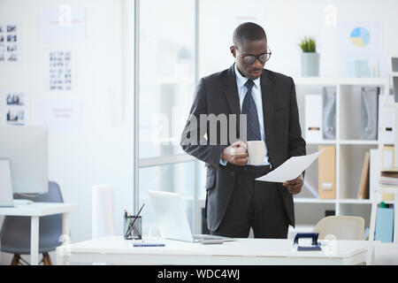 Grave uomo africana in occhiali e in abito nero in piedi vicino al suo luogo di lavoro la lettura di un documento e di bere il caffè Foto Stock