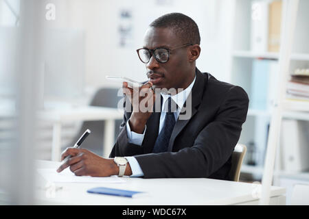 Giovani manager africani in abbigliamento formale tramite telefono cellulare e invio di un messaggio audio mentre si lavora al tavolo in ufficio Foto Stock