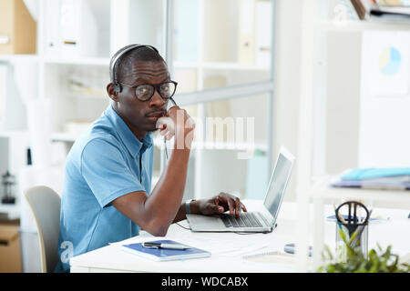 Ritratto di giovane designer africani che indossa le cuffie e occhiali seduto al suo posto di lavoro nella parte anteriore del computer portatile e guardando la telecamera in ufficio Foto Stock