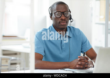 Ritratto di giovane imprenditore africano in camicia che indossano le cuffie seduto al suo posto di lavoro e guardando la fotocamera Foto Stock