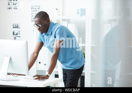 African giovane designer in occhiali e casual in piedi vicino al tavolo e guardando il monitor del computer in ufficio Foto Stock